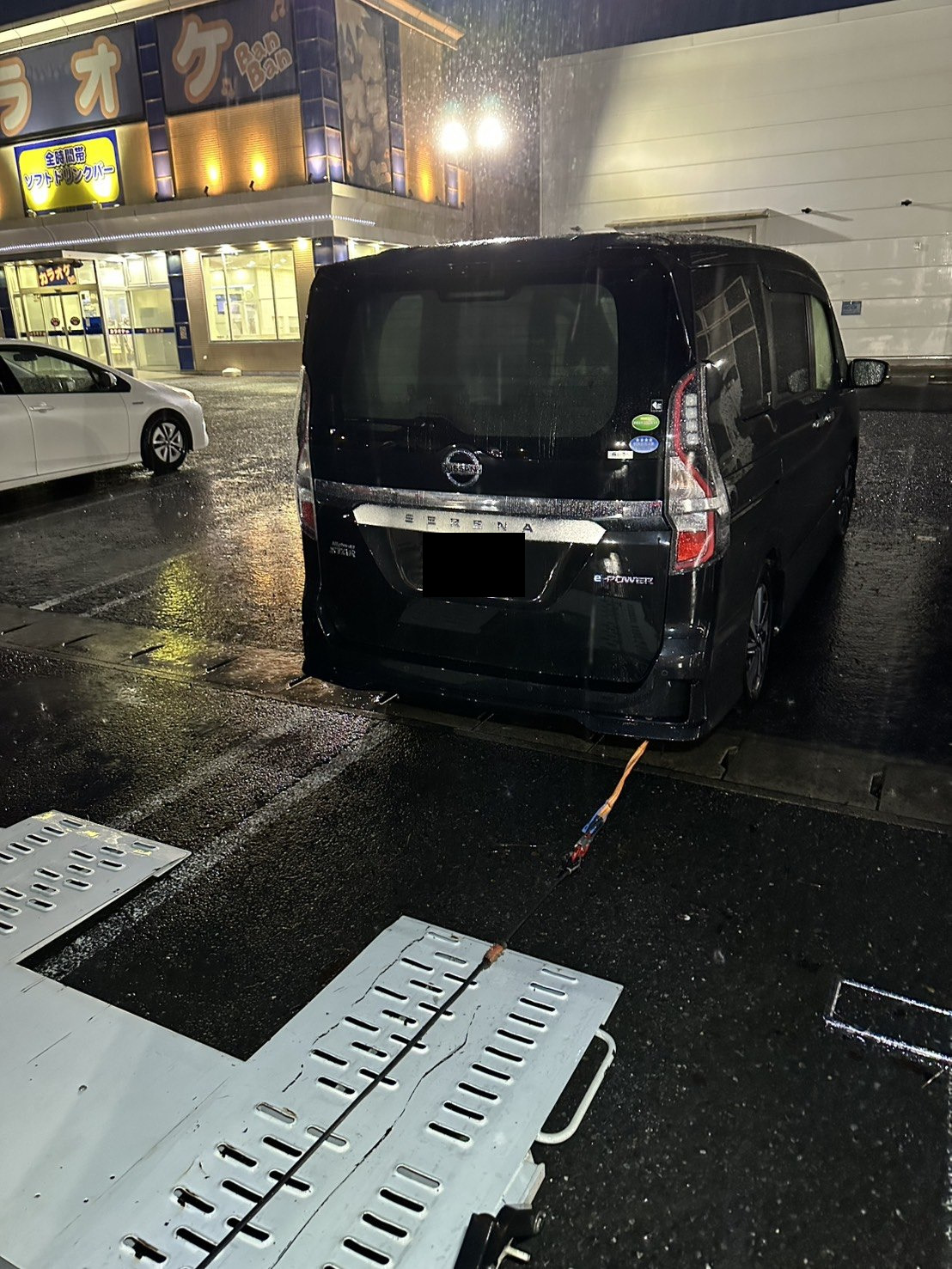 強い雨が降る夜の駐車場で、積載車のラダーを敷いて黒いミニバンの積み込み準備をしている様子