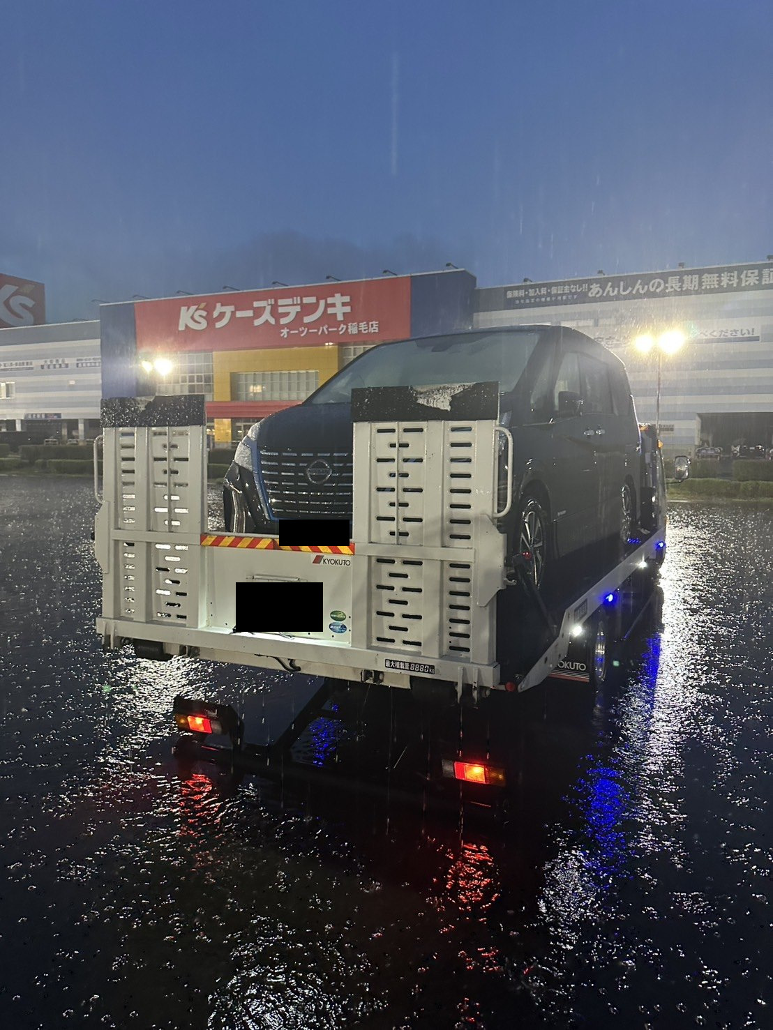 雨の降る駐車場で、積載車の荷台に載せられた黒いミニバンを後方から見た様子