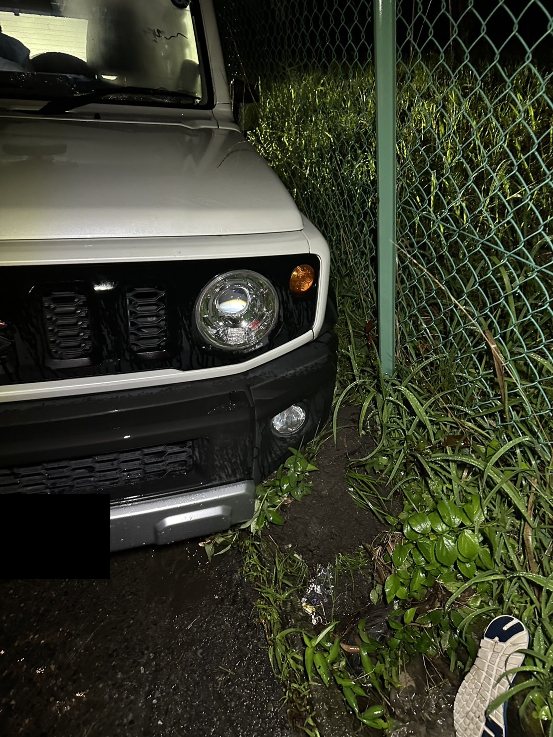 夜間の雨の中、フェンス沿いの路肩に前輪を落とした状態で停車している白いSUVの前部