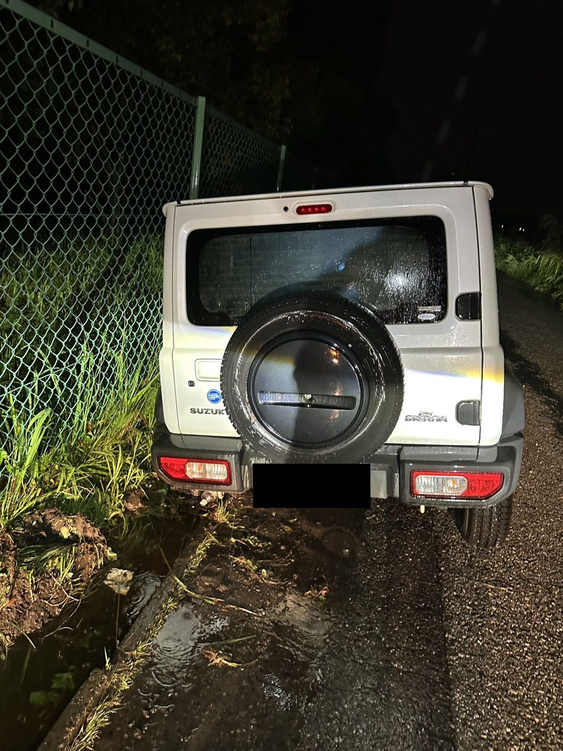 夜間の雨の中、道路脇の側溝側に左のタイヤを落として傾いている白いSUVの後部