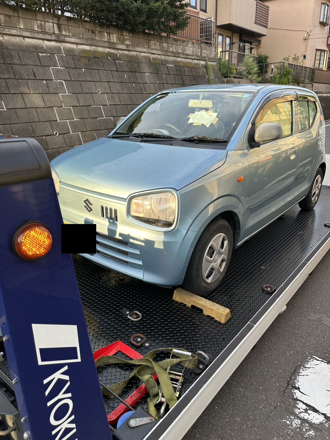積載車の荷台に載せられた水色の軽自動車を前方から見た様子