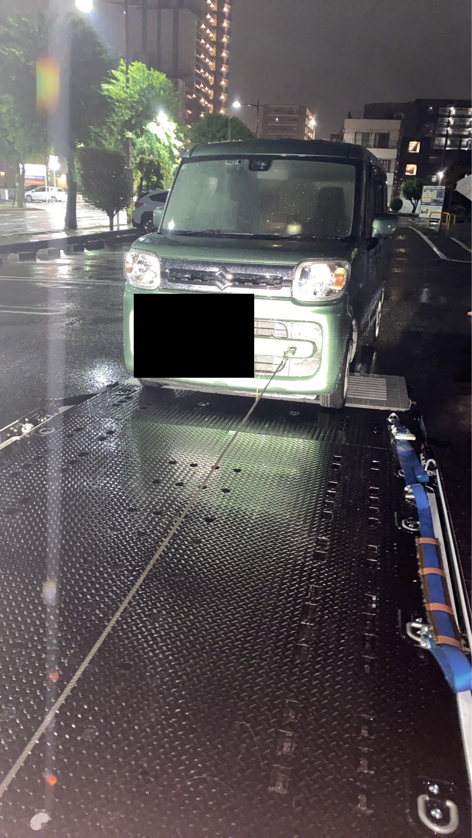 雨の降る夜間、積載車の荷台に載せられた緑色の軽自動車を正面から見た様子
