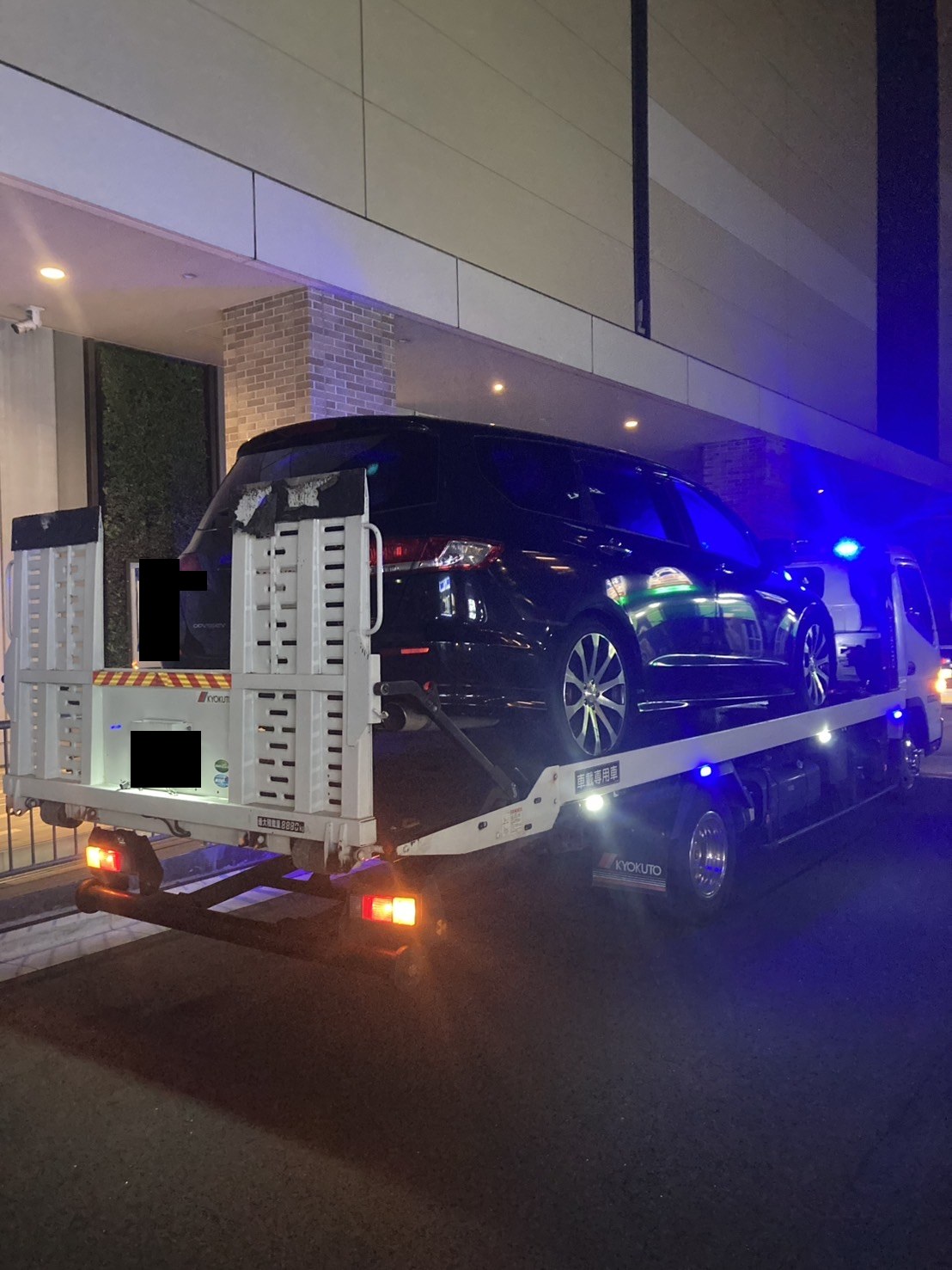 夜間、積載車上の黒いミニバンを斜め後方から見た様子（ナンバープレートはマスク処理）