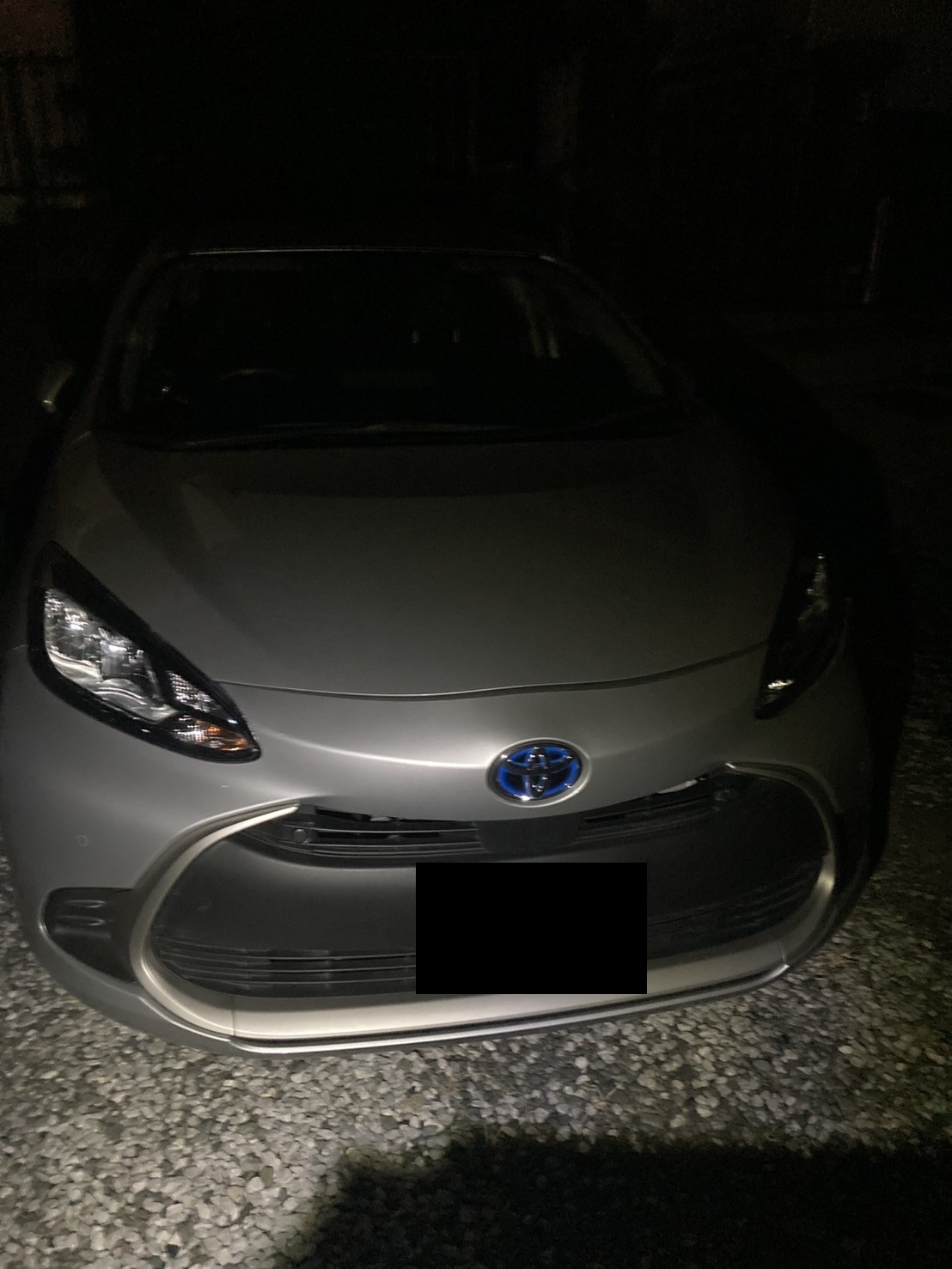 夜間、砂利敷きの場所に停車したシルバーのコンパクトカーを正面から見た様子（ナンバープレートはマスク処理）
