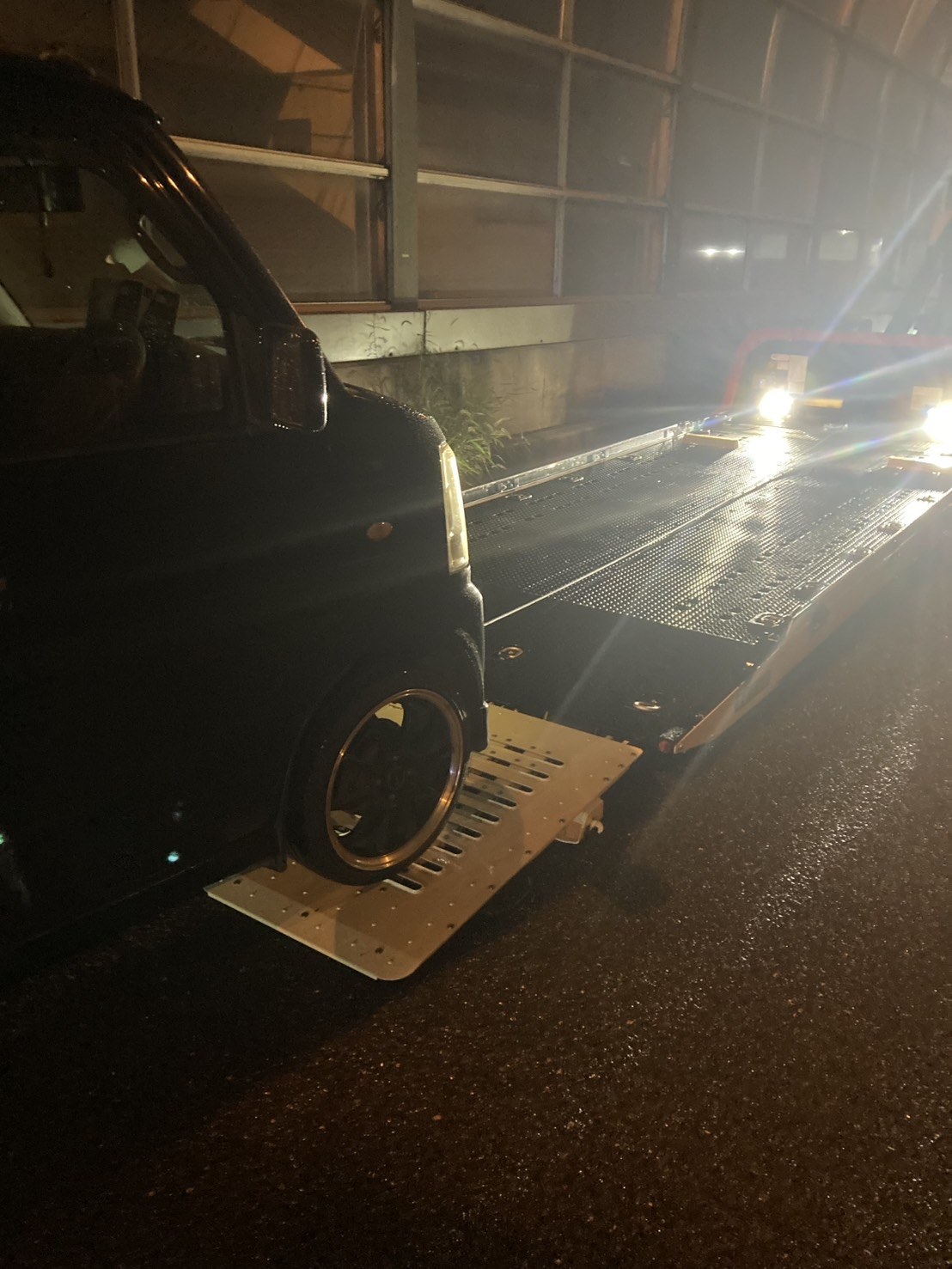 夜間、防音壁のある道路で積載車のスロープに前輪を載せた軽ワゴンを側方から見た様子