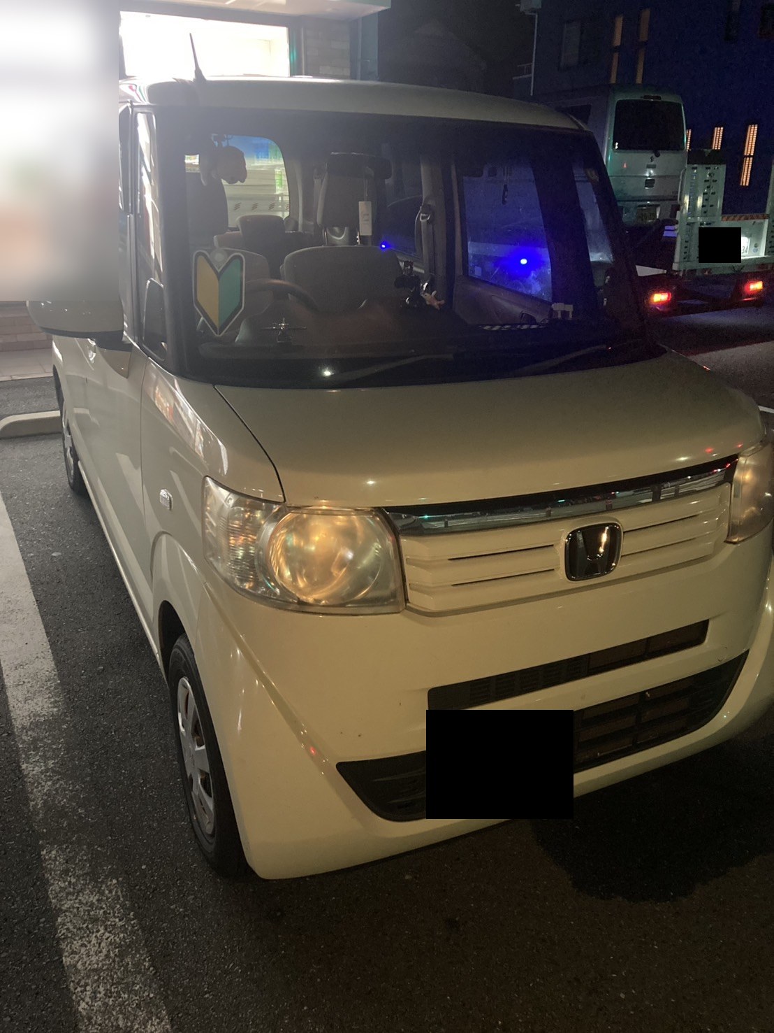 夜間、駐車スペースに停車した白い軽自動車を前方から見た様子（ナンバープレートはマスク処理）